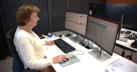 Mevrouw zit aan bureau met 2 schermen in de achtergrond staat een zwarte kast